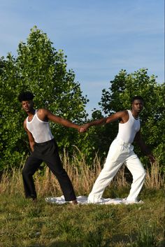 two young men holding hands while standing on a blanket in the grass with trees behind them