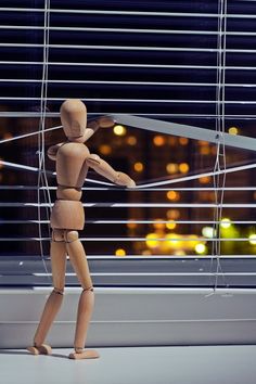 a wooden mannequin standing on a window sill in front of blinds with city lights behind it