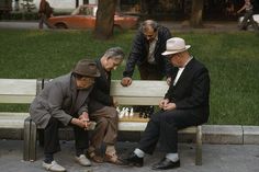 three older men playing chess on a park bench in the middle of an urban area