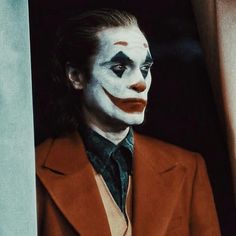 a man dressed as the joker looks out from behind a curtain with his face painted red and white