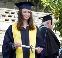 a young woman in graduation gown holding a stick and smiling at the camera while other people look on