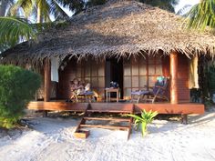 two people sitting on chairs in front of a hut with thatched roof and palm trees