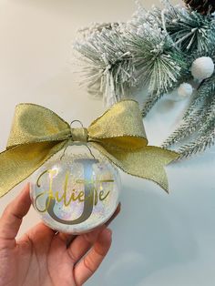 someone holding up a personalized glass ornament with a gold bow on it