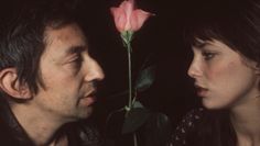 a man standing next to a woman with a pink rose in her hand and looking at him