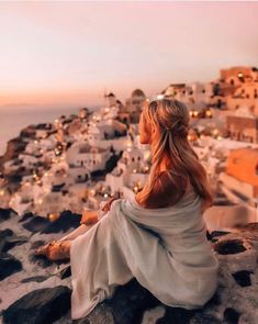 a woman sitting on top of a rock next to the ocean at sunset with lights in the background