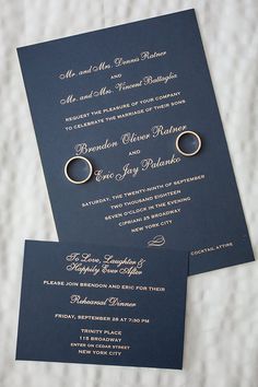 two wedding rings are on top of the navy blue and gold wedding stationery, which is laid out in front of each other