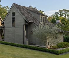 a gray house with hedges and trees in the front yard, surrounded by green grass