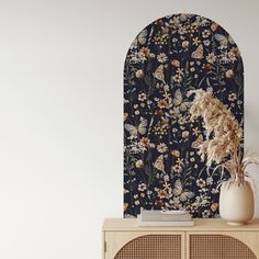 a white vase sitting on top of a wooden cabinet next to a wall with flowers