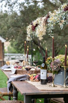 an outdoor table with candles, flowers and bottles of wine on it in the grass