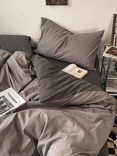 an unmade bed with grey sheets and pillow cases next to a lamp on a nightstand