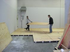 two men in an unfinished room with plywood boards on the floor and one man holding a piece of wood