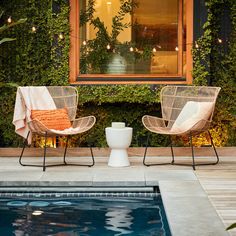 two chairs sitting next to a swimming pool in front of a house with plants growing on the wall