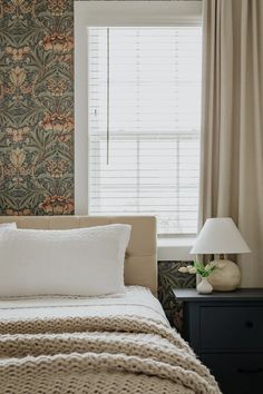 a bed sitting under a window next to a night stand with a lamp on it