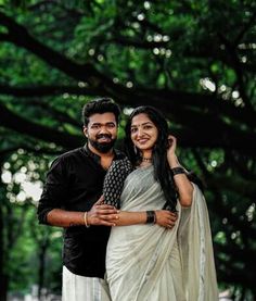 a man and woman standing next to each other in front of some trees with their arms around each other