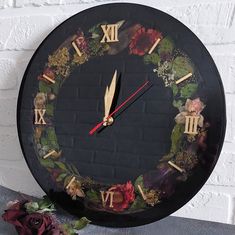 a black clock with flowers and roman numerals on the face next to a brick wall