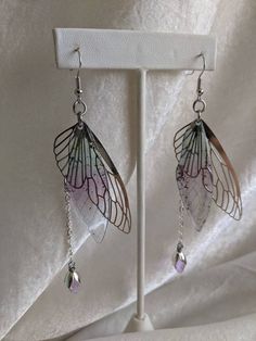 These gorgeous and delicate fairy earrings feature metal and resin wings with a hanging drop crystal. Perfect for special occasions or to add a bit of magic to your every day! The primary color refers to the metal The secondary color refers to the resin wing Silver Fairy Earrings For Party, Silver Fairy Style Party Earrings, Fairycore Silver Earrings For Gift, Silver Fairy Dangle Earrings, Silver Fairy Style Dangle Earrings, Fairy Style Silver Earrings, Silver Fairycore Drop Earrings, Silver Fairy Grunge Dangle Jewelry, Fairy Grunge Silver Dangle Jewelry