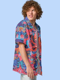 a young man with curly hair wearing a colorful shirt and white pants, standing in front of a blue background