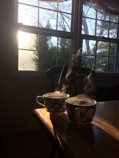 two coffee mugs sitting on top of a wooden table next to a large window