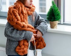 a woman standing next to a window with a large scarf on her shoulders and a small potted tree in the background