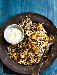 a plate filled with rice and vegetables next to a bowl of yogurt on a blue wooden table