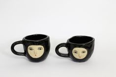 two black mugs with faces painted on them sitting next to each other in front of a white background