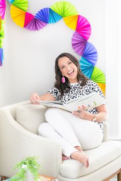 a woman sitting on a couch holding a book and looking at the camera with colorful paper decorations behind her