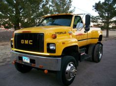 a large yellow truck parked in a parking lot next to some trees and dirt ground