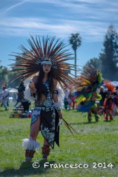 a woman dressed in native american clothing and headdress walking through the grass with feathers on her head