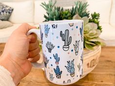 a person holding a coffee mug with cactus designs on it and succulents in the background