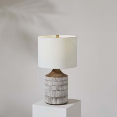 a table lamp with a white shade sitting on top of a small square block in front of a gray wall