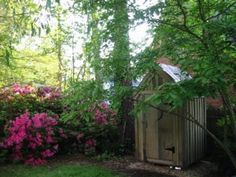 an outhouse in the middle of some bushes and trees with pink flowers around it