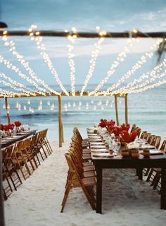 tables set up on the beach with lights strung over them and red flowers in vases