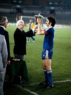 an old man holding up a trophy while standing next to another man on a soccer field