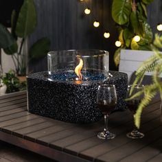 a lit candle is sitting on a table with two wine glasses and potted plants in the background