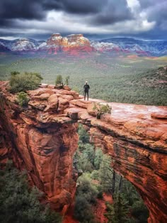 a man standing on the edge of a cliff looking down at trees and mountains under a cloudy sky