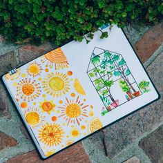 an open notebook with drawings on it sitting next to some green plants and flowers in the background