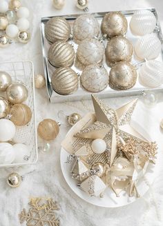 a table topped with white and gold christmas ornaments next to silver trays filled with baubles