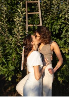 two women kissing each other in front of trees and an old ladder with leaves on it