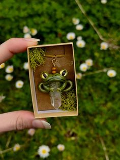 a person holding up a tiny frog in a box with moss on it's face