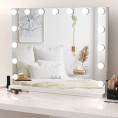 a white bed sitting under a large mirror with lights on it's sides and pillows in front of it