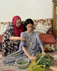 two women are sitting on the floor and one is cutting up some vegetables while the other looks at her phone