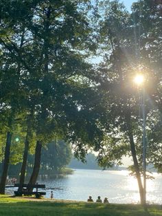 the sun shines brightly through some trees near a lake with people sitting on benches