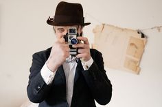 a man in a suit and hat taking a selfie with his camera while standing in front of a bed