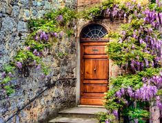 an old stone building with purple flowers growing on it's side and a wooden door