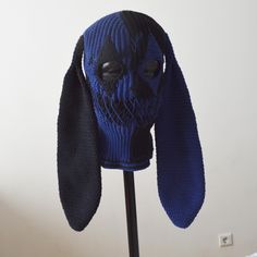 a blue knitted dog mask on a black head stand in front of a white wall