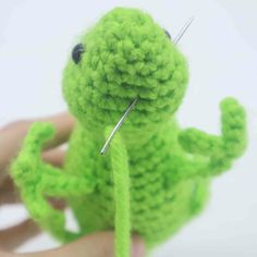 a green crocheted frog sitting on top of a person's hand with needles in its mouth