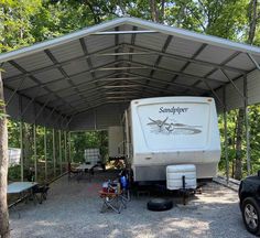 an rv is parked under a metal carport in the woods with tables and chairs around it