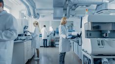 two women in white lab coats are looking at computer screens and printer equipment on the counter