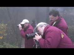 two men are taking pictures in the foggy woods with their camera phones and one man is wearing a red jacket