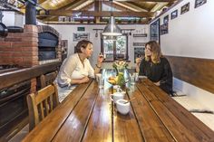 two women sitting at a wooden table with food in front of them and brick oven behind them